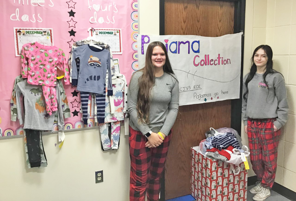 Two female early childhood education students stand next to their box filled with donated pajamas